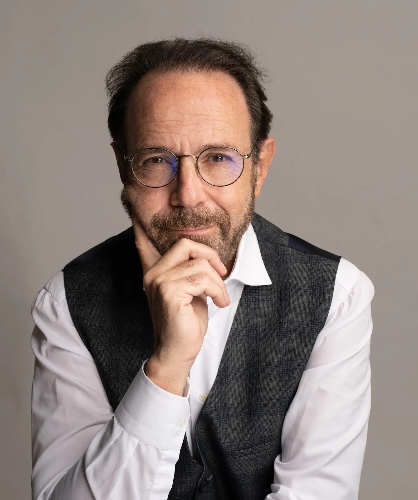 Marc Levy, French bestselling novelist, wearing round glasses, a white shirt and dark waistcoat, resting his chin on his hand.